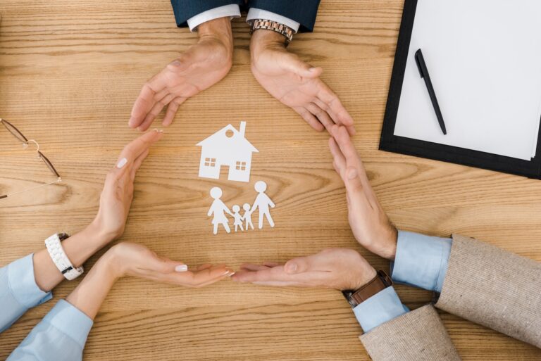 People making circle with hands on wooden table with paper house and family inside, life insurance