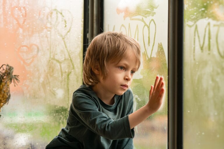 A little boy painting in children`s handwriting on wet glass of a window in the light of city lights